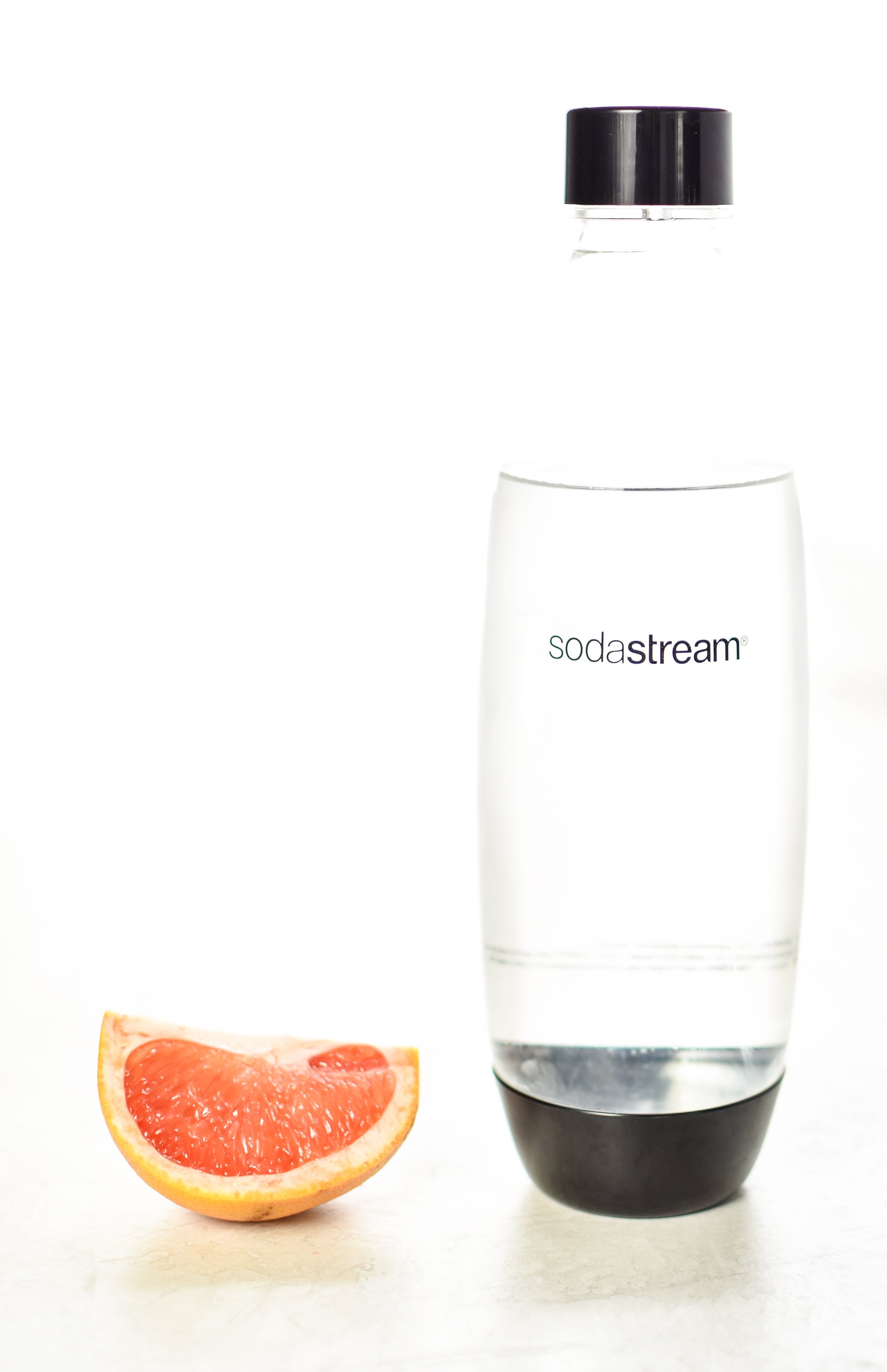 A slice of grapefruit sitting next to a SodaStream carbonating bottle filled with cold water in a bright room.