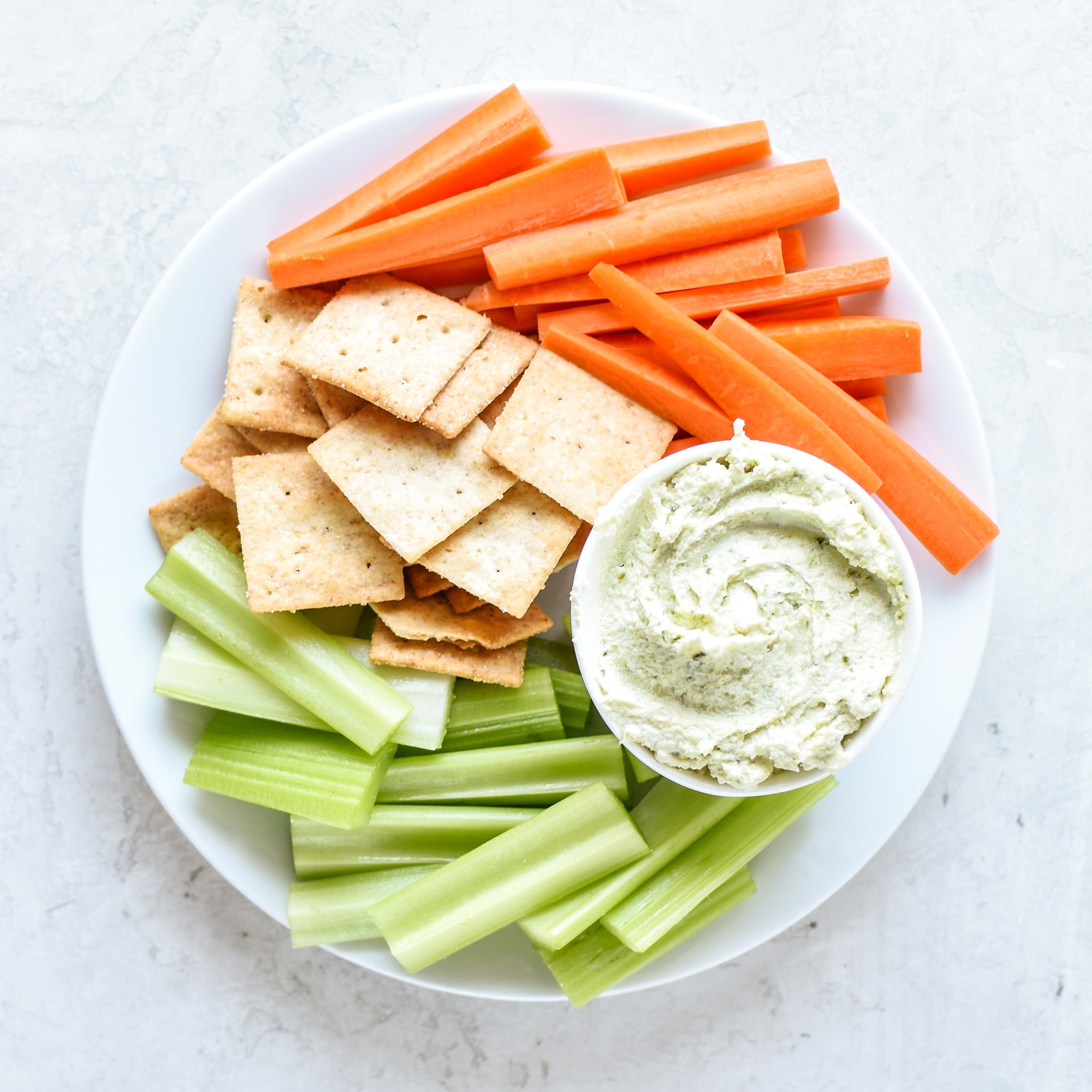 A bowl of 3 Ingredient Pesto Goat Cheese Dip with carrots, crackers and celery.