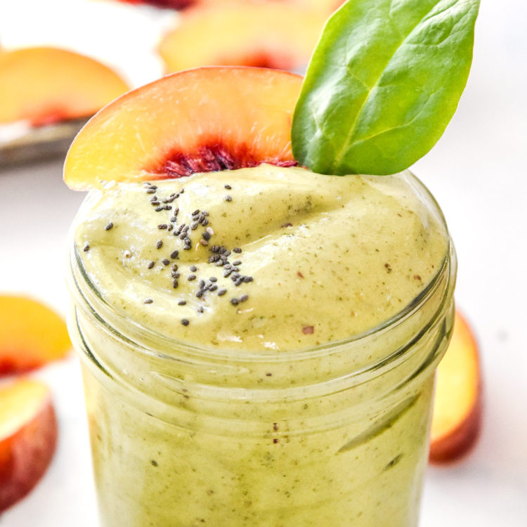 peaches and green protein smoothie with spinach leaf, chia seeds and peach slice on top.