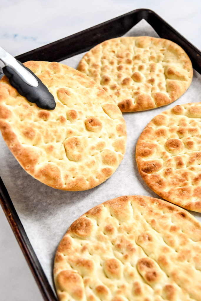 pitas toasted on a rimmed sheet pan, one being held with tongs.