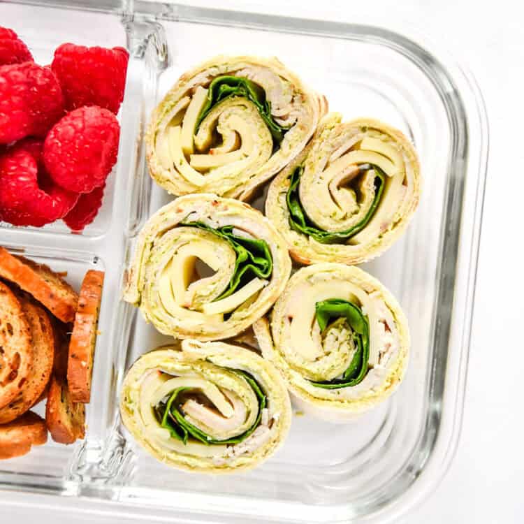 turkey pesto pinwheels in a glass meal prep container with raspberries and bagel chips.